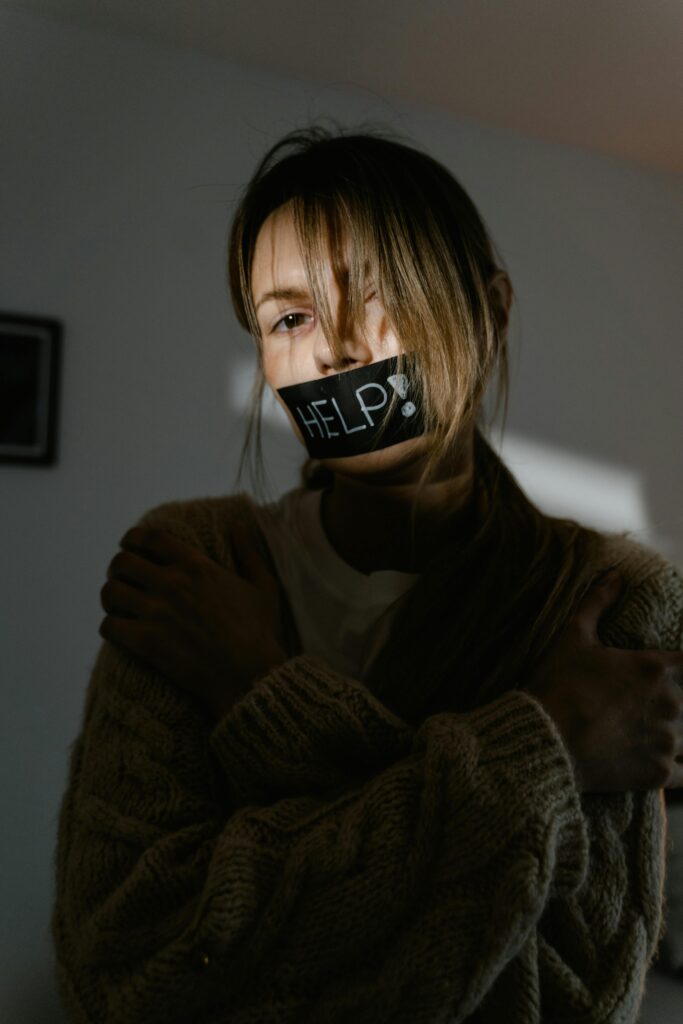 A woman with 'help' tape over mouth, expressing distress and urgency indoors.